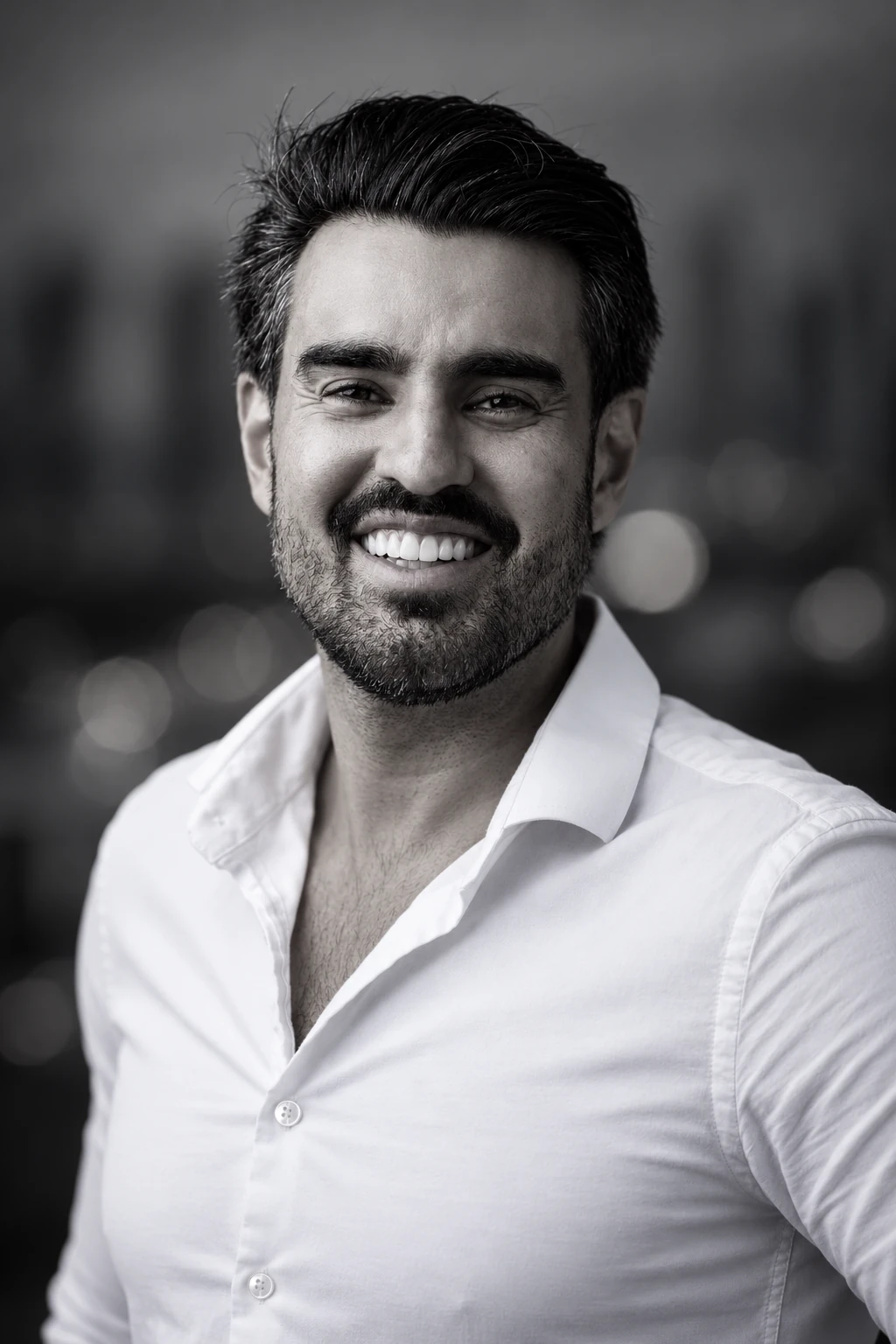 Retrato em preto e branco de Bruno Lima, sorrindo, de camisa branca, com a cidade desfocada ao fundo.